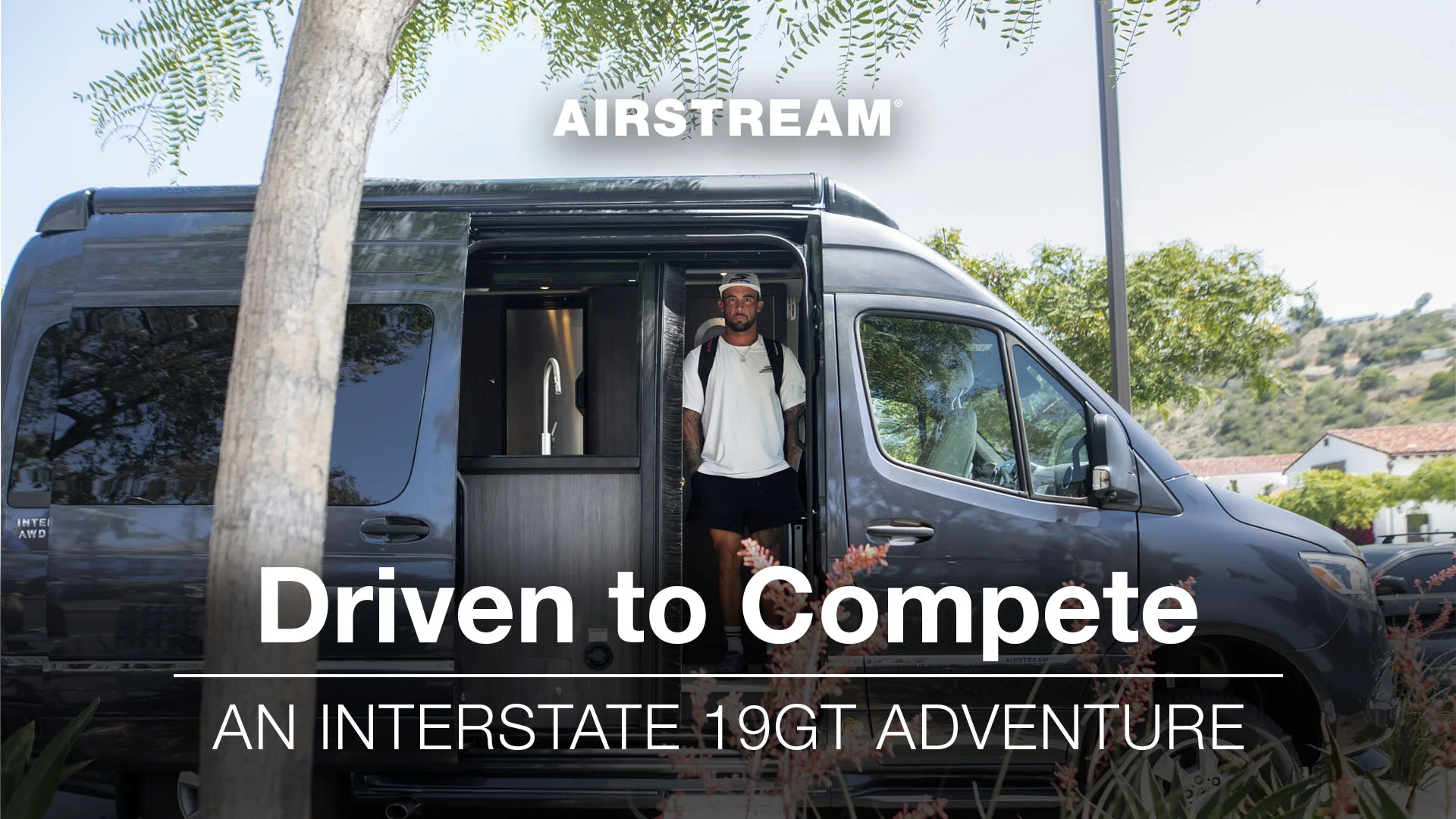 Man stands in the doorway of a black Airstream Interstate 19GT camper van, surrounded by trees and greenery. Text: "Driven to Compete."