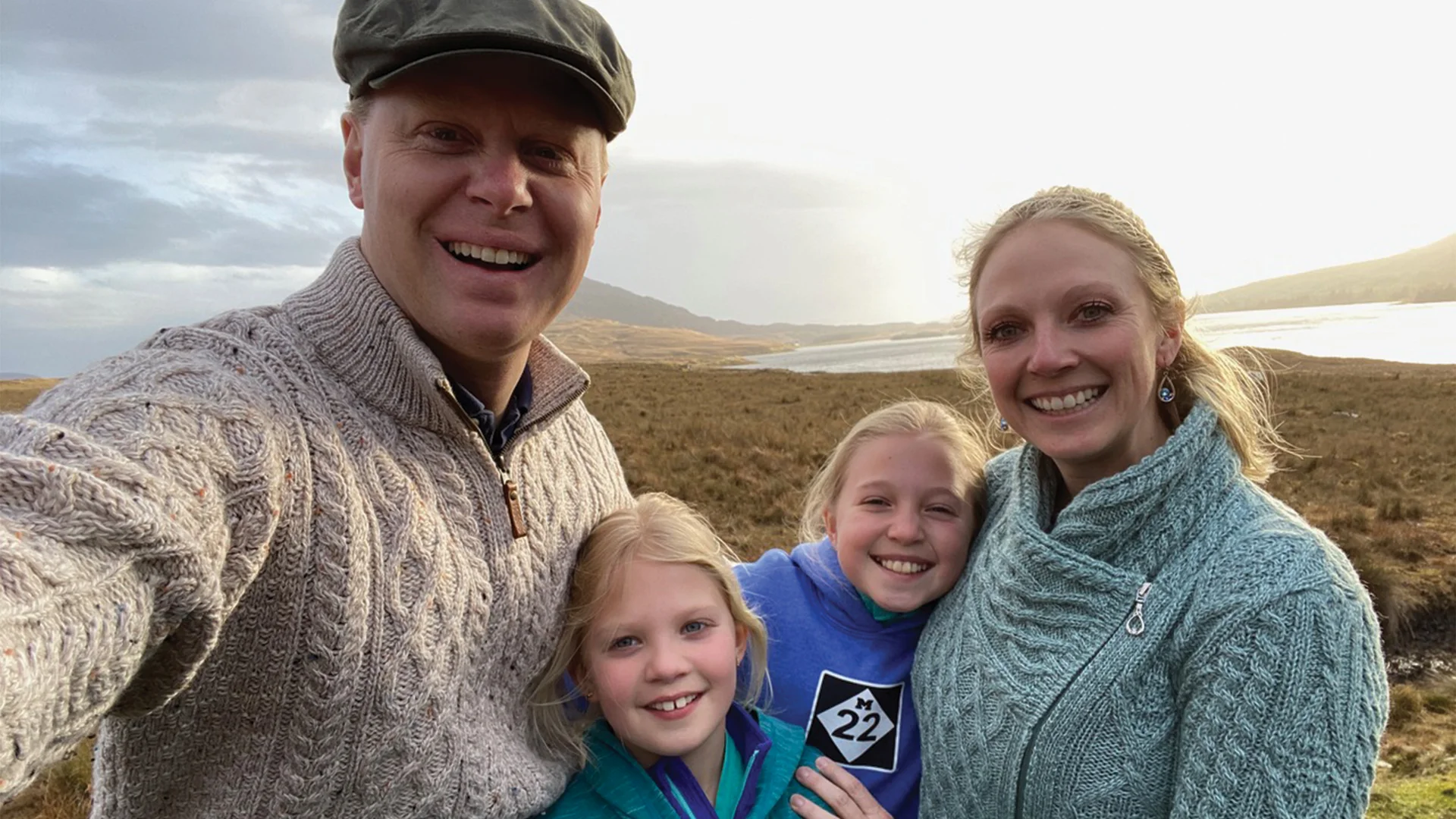 A smiling family of four takes a selfie in a scenic outdoor setting, with open fields, a distant river, and cloudy skies in the background.