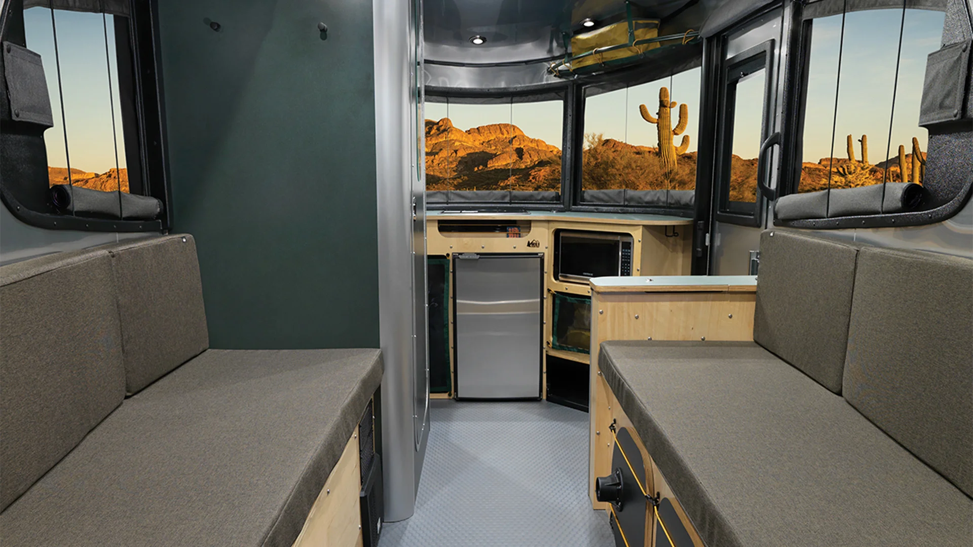 Interior of a modern RV with gray seating, a compact kitchen area, and large windows showcasing a desert landscape with cacti at sunset.