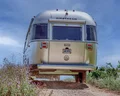A silver Airstream trailer drives down a dirt road surrounded by grass and wildflowers under a blue sky, with a license plate reading “LIVE RWD.”.