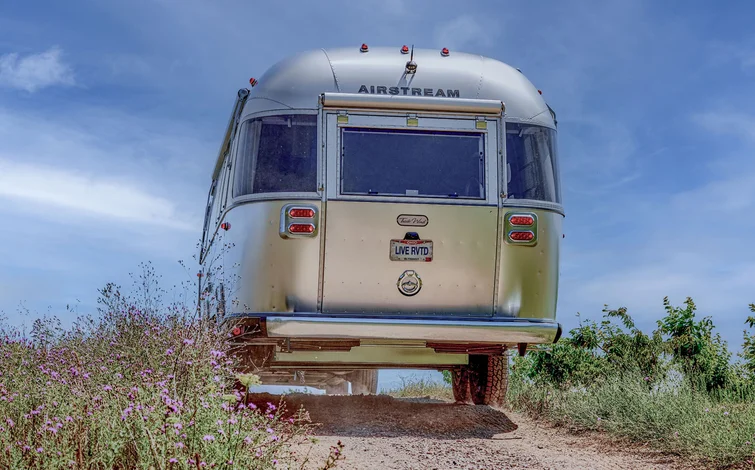 A silver Airstream trailer drives down a dirt road surrounded by grass and wildflowers under a blue sky, with a license plate reading “LIVE RWD.”.
