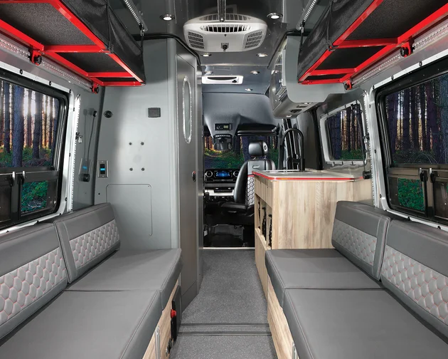 Interior of a modern camper van with gray cushioned benches on both sides, wood accents, large windows showing a forest view, and overhead red storage racks. The driver’s area is visible at the front.