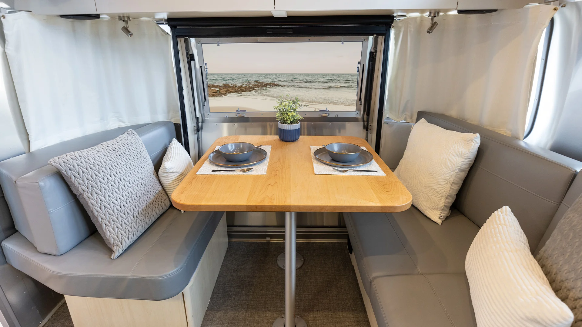 Modern RV dining area with a wooden table set for two, grey cushioned benches, and a window view of the ocean.