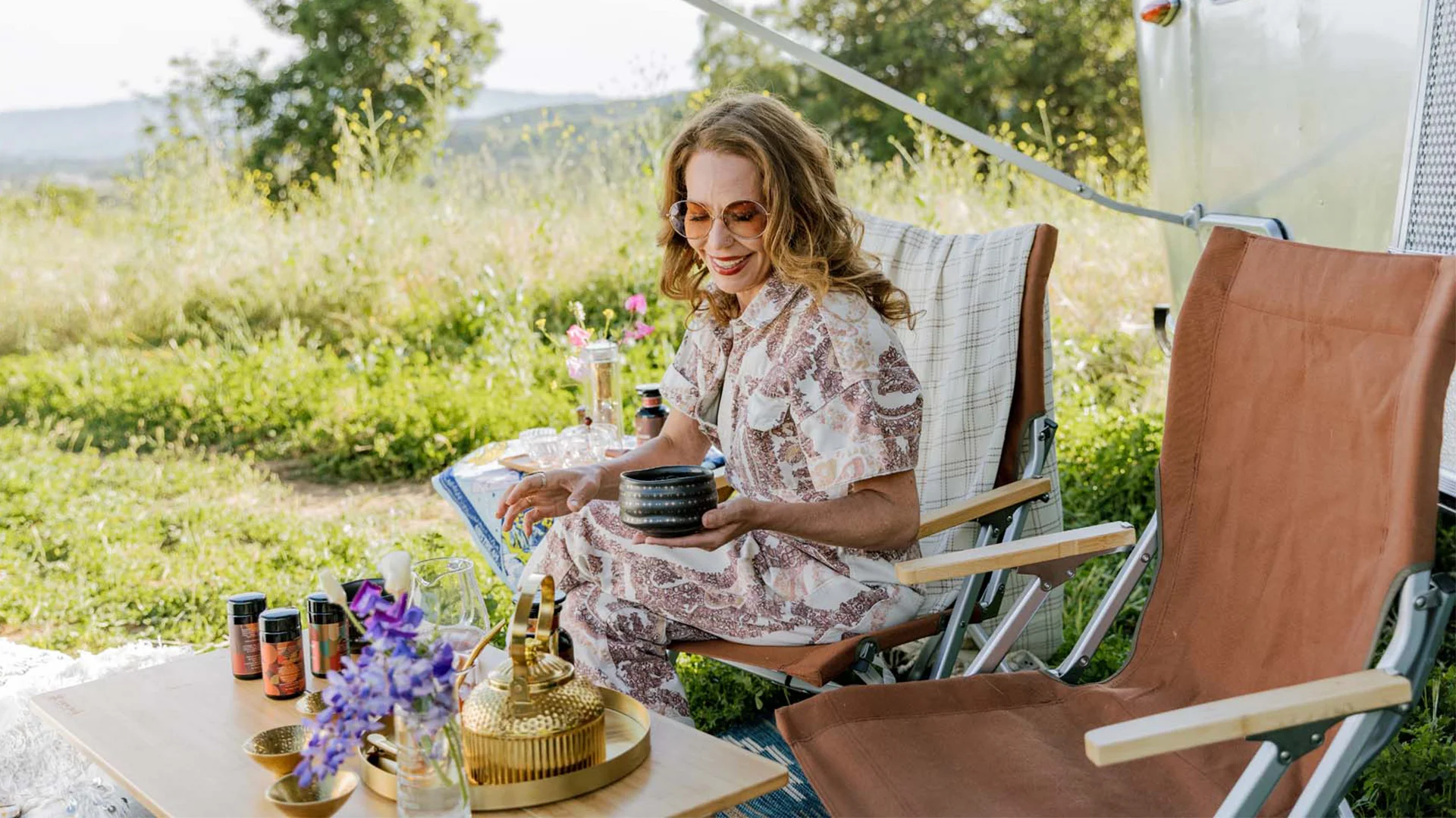 Woman in a patterned dress sits on a chair outdoors, holding a small bowl. Table with flowers, jars, and a teapot nearby. Sunny day.