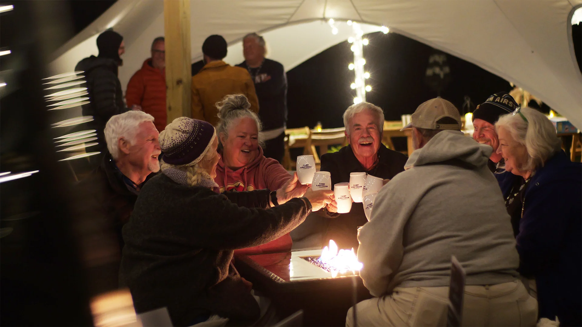 Group of people laughing and toasting with mugs around a fire pit under string lights at night.