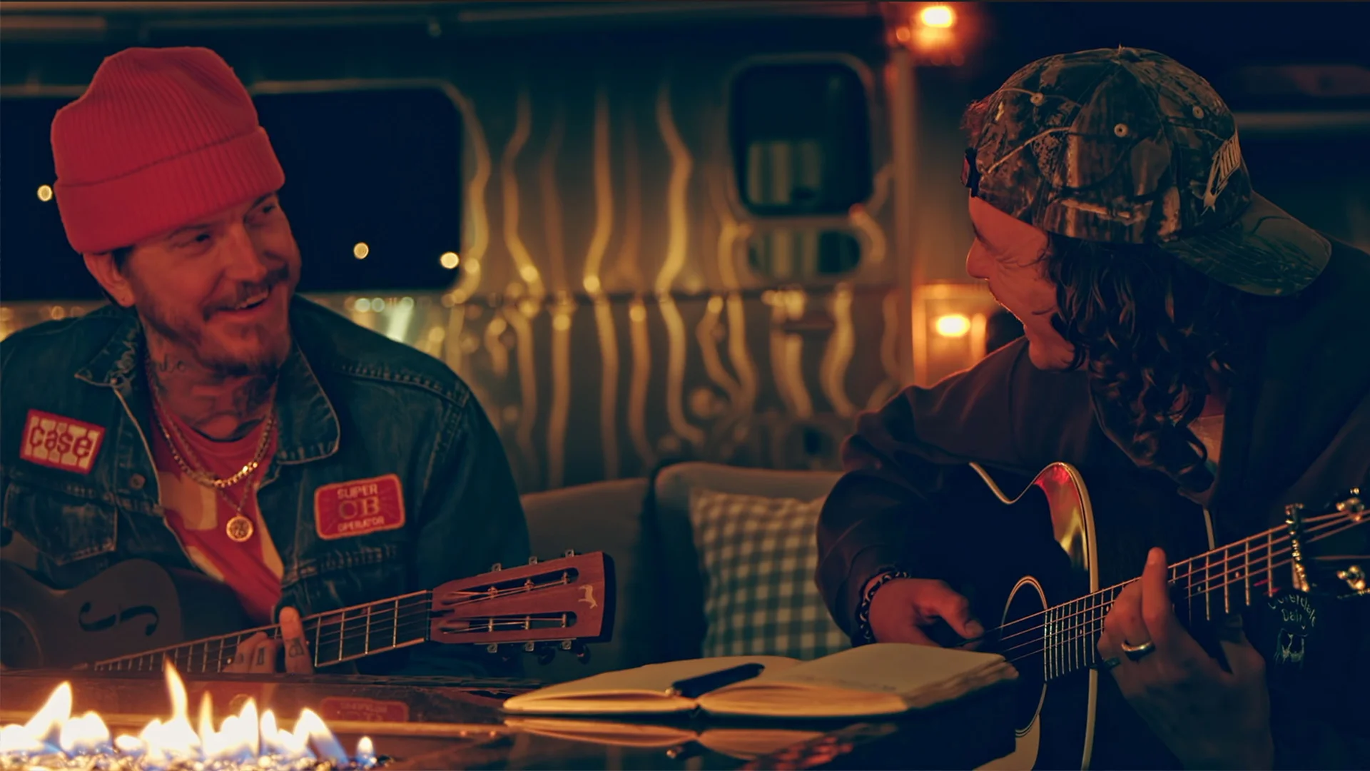 Two men playing guitars around a fire pit, smiling at each other, with a notebook and pen on the table, in a cozy outdoor setting in front of an Airstream travel trailer..