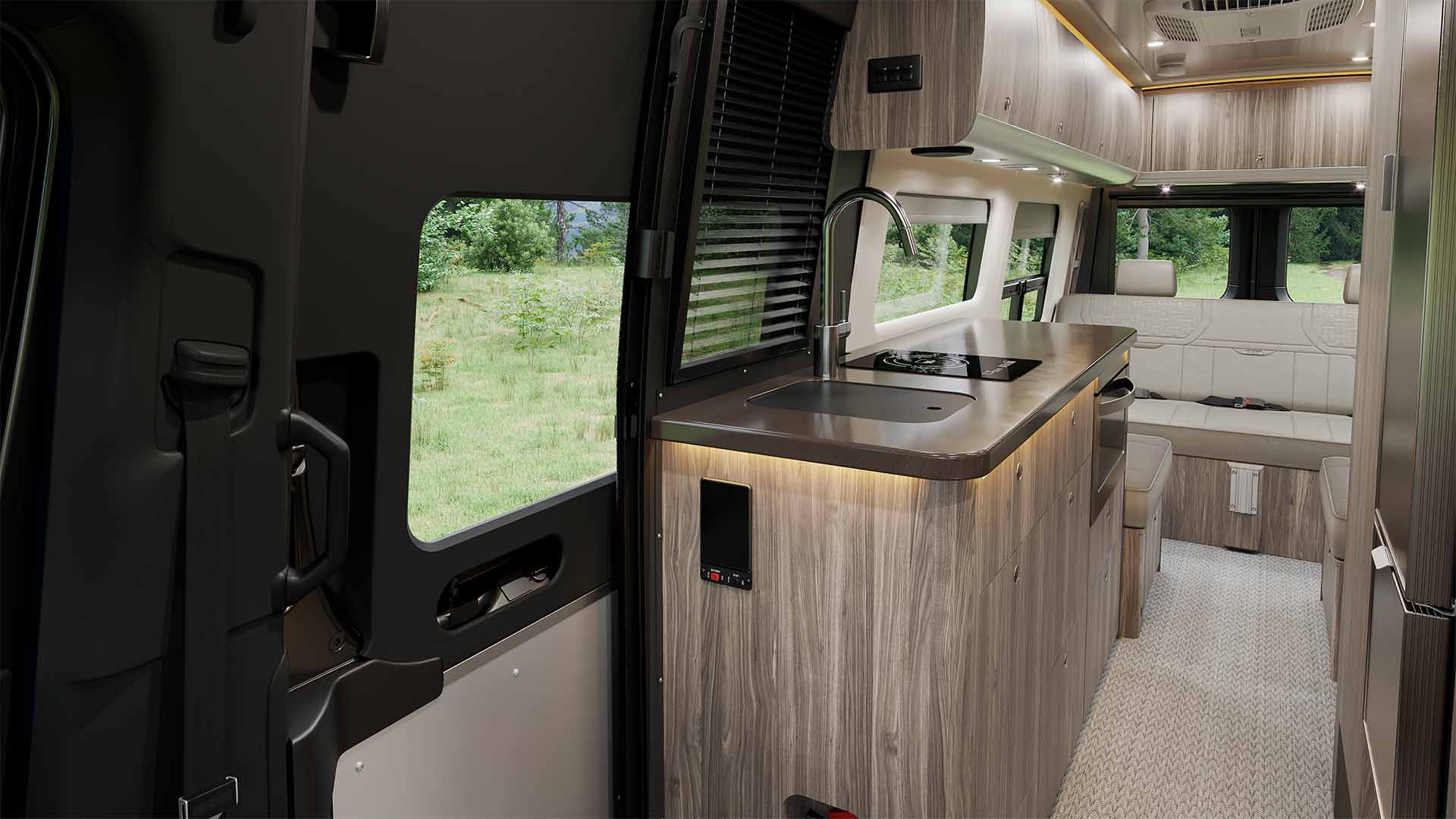 Interior of a camper van with a modern kitchenette, featuring a sink, stove, and wood cabinets, leading to a cozy seating area.