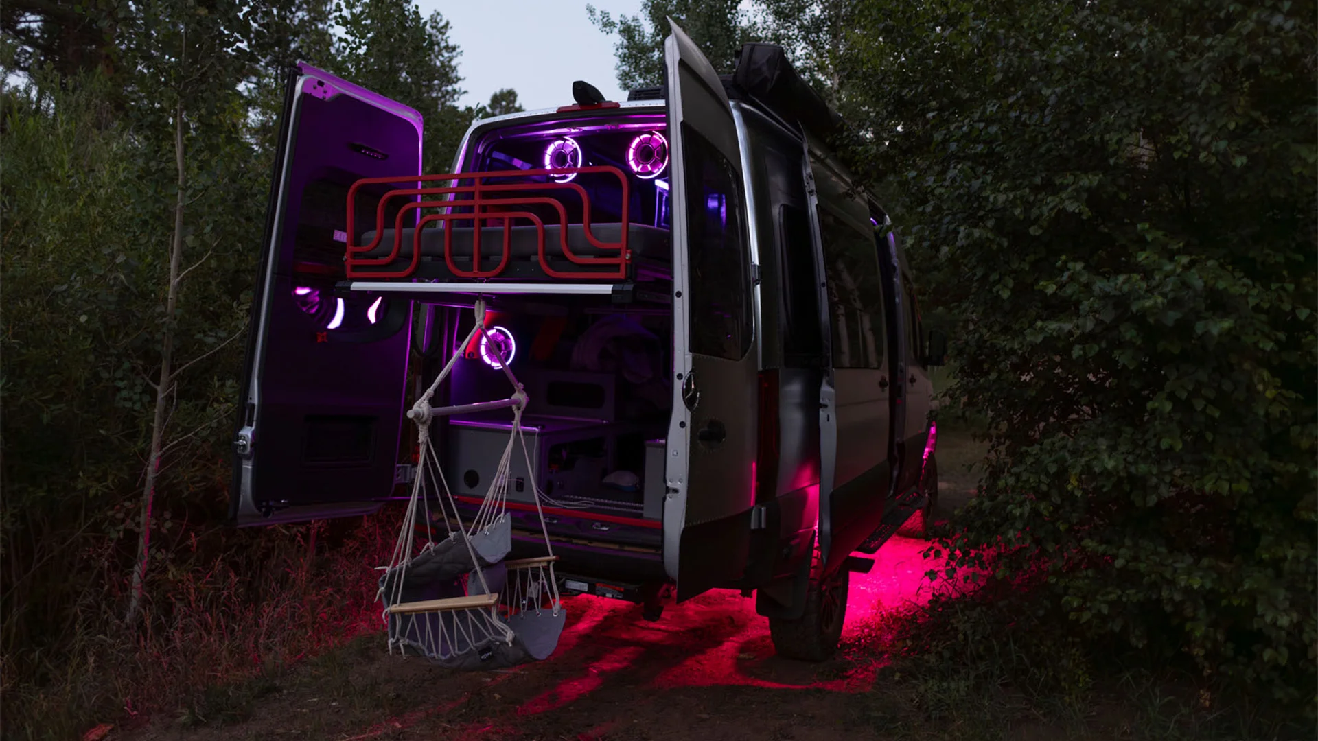 A van with open doors and neon pink lights illuminates a wooded area. Inside, spears and a hammock are visible.