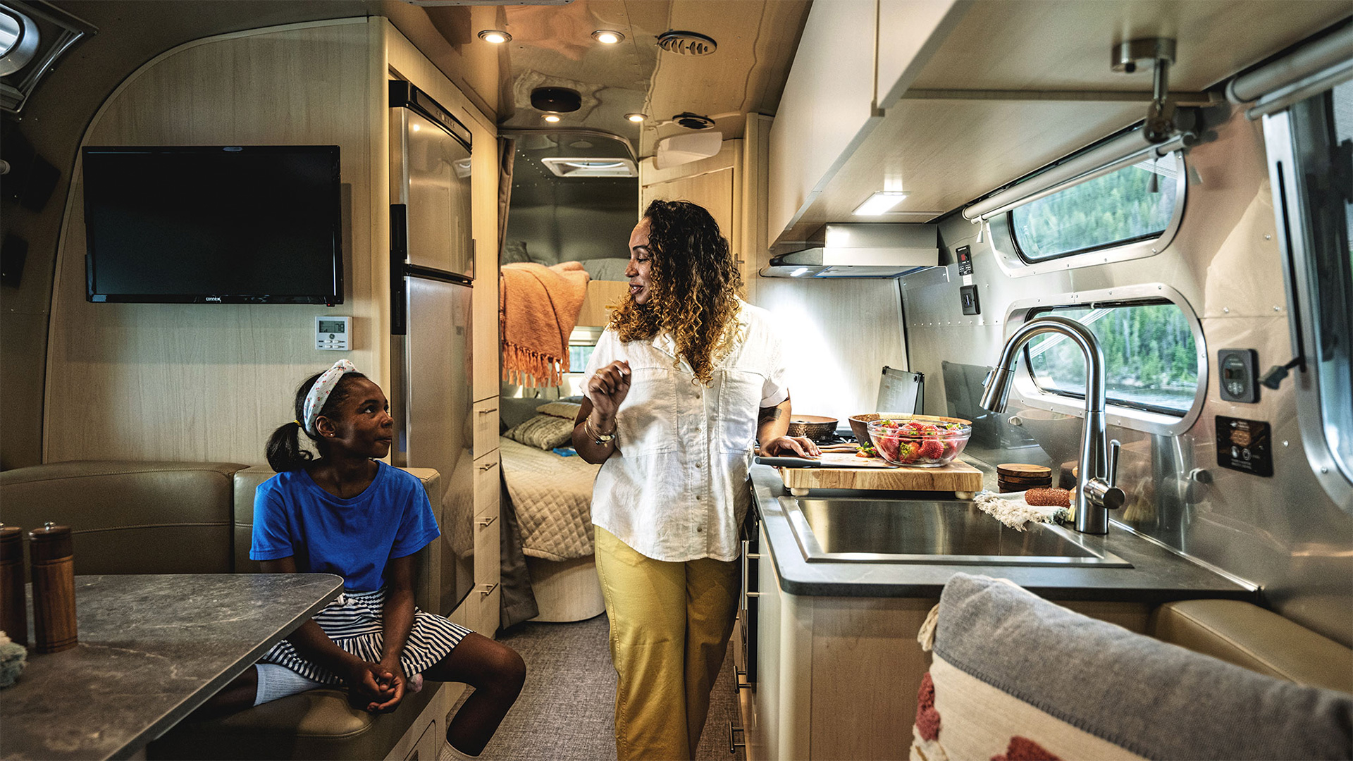 Woman and girl in an RV kitchen; the woman is preparing food while the girl sits nearby, smiling. Cozy and well-lit interior.