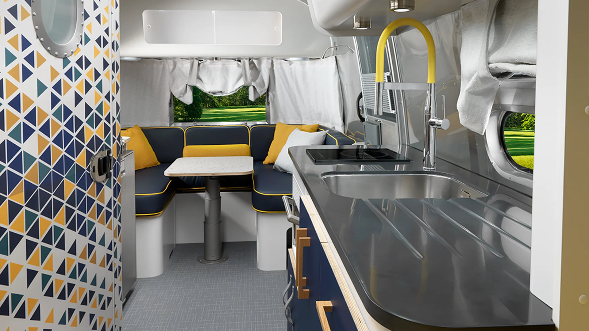 Modern RV interior with a sleek kitchen on the right, featuring a metallic counter and yellow-accented faucet. In the back, a dining nook with navy, yellow, and white cushions surrounds a small table. Geometric pattern on the door.