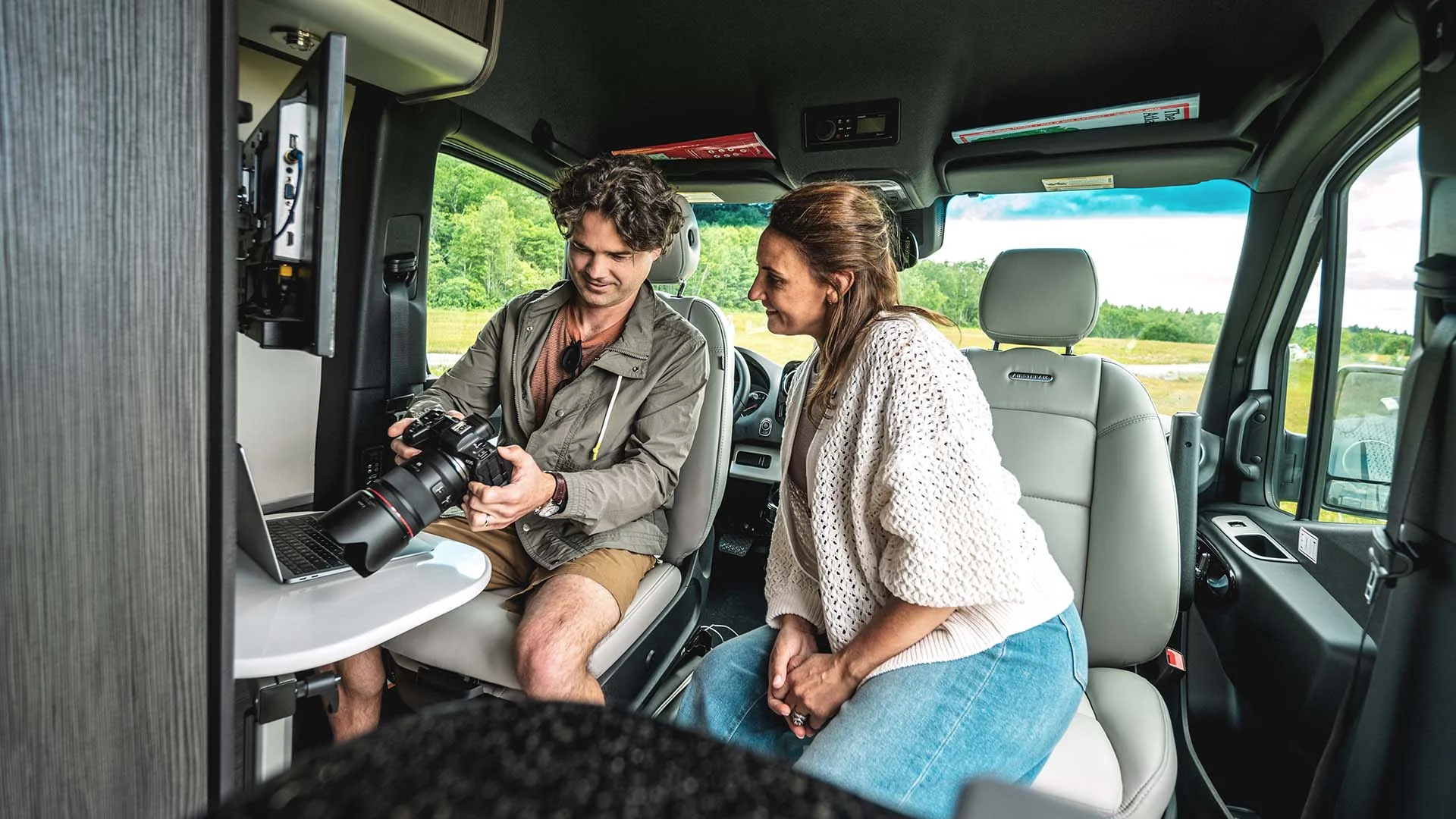Two people sit inside a van, examining a camera next to a laptop on a small table, with a scenic view outside.