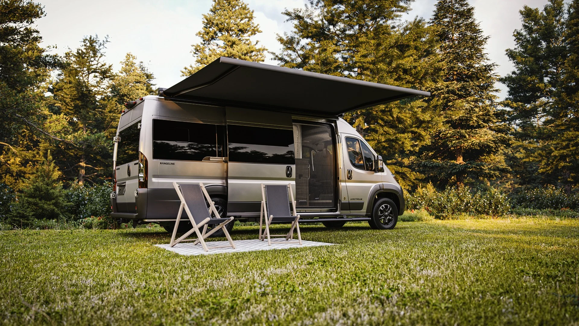 Camper van with extended awning parked on grass, two chairs in front, surrounded by tall trees on a sunny day.