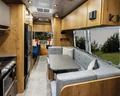 Modern RV interior with wooden cabinets, gray seating around a table, and a kitchen area; large window overlooking greenery.