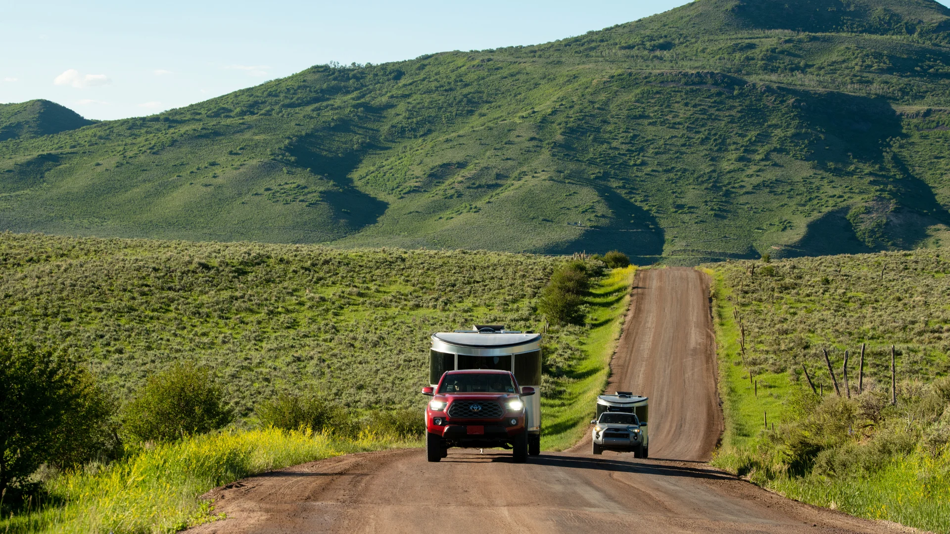 Red truck towing a trailer on a dirt road in a lush green hilly landscape, with another vehicle following behind.