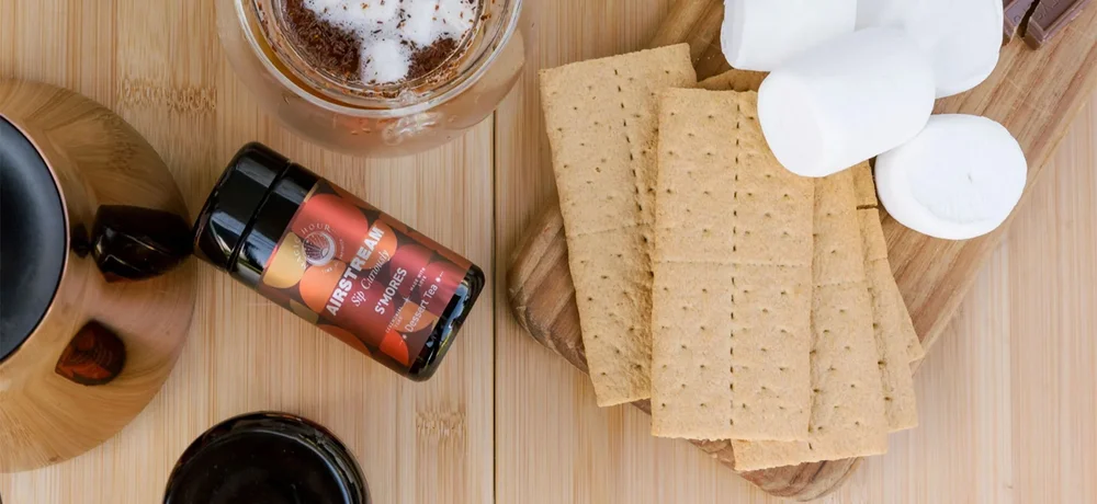 S'mores ingredients: marshmallows, graham crackers, and chocolate on a wooden board with a spice jar and a pot of coffee nearby.