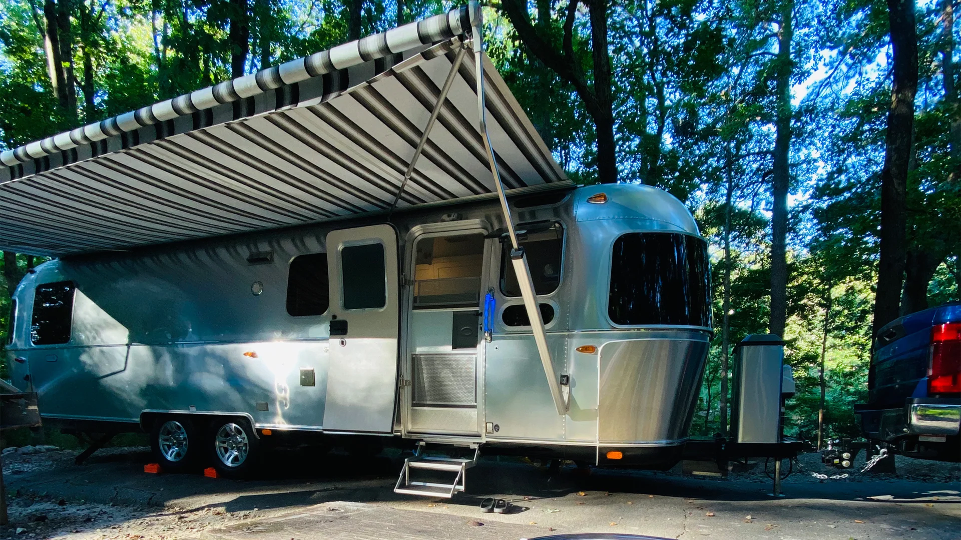 A silver Airstream travel trailer with an extended striped awning is parked on a paved campsite surrounded by tall trees and dappled sunlight. A blue pickup truck is partially visible on the right.