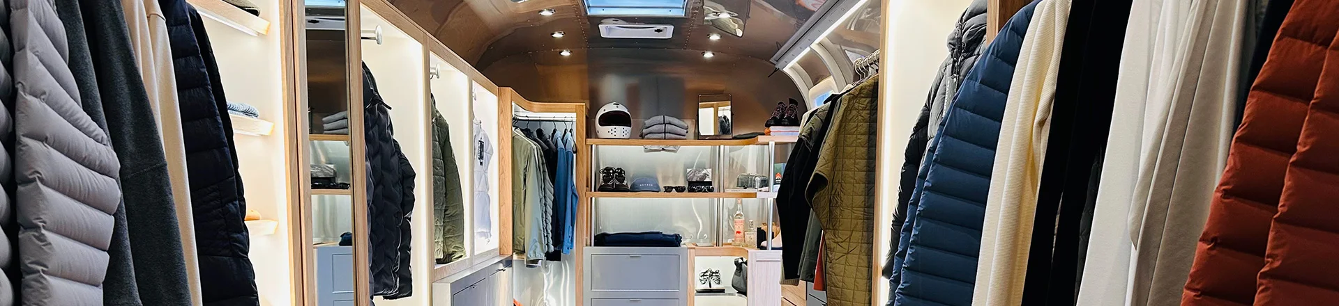 Interior of a mobile clothing boutique in an Airstream, with racks of jackets and shirts, shelves with hats and accessories, and a wooden floor.
