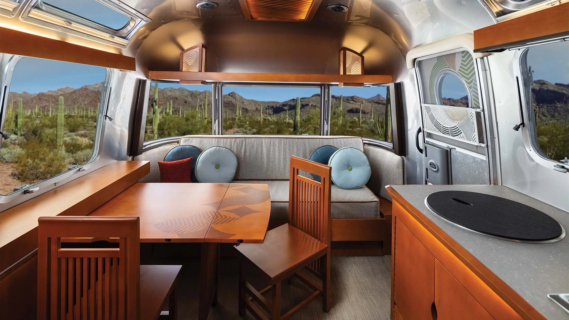 Interior of a modern Airstream trailer featuring wood furnishings, a dining area, and large windows overlooking a desert landscape with cacti.