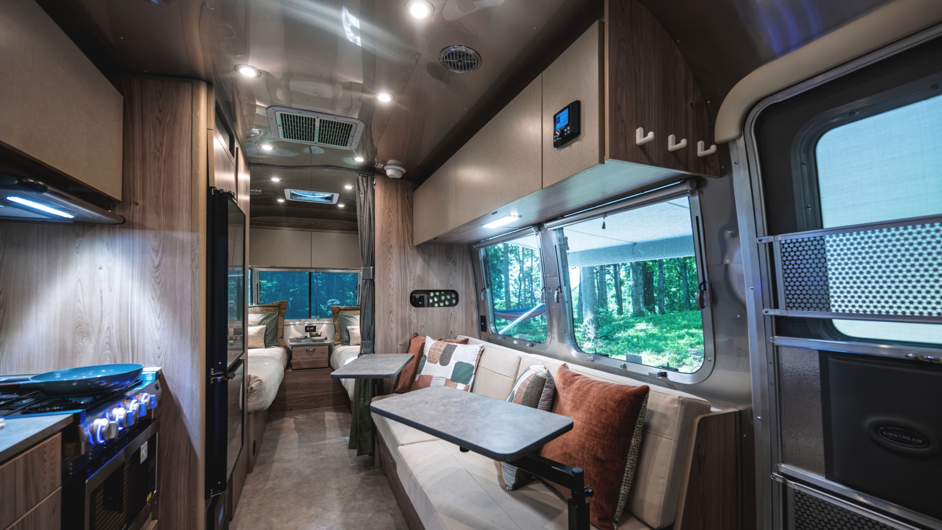Interior of a sleek RV featuring a modern kitchen, cozy seating area with cushions, and a window view of lush green trees outside.