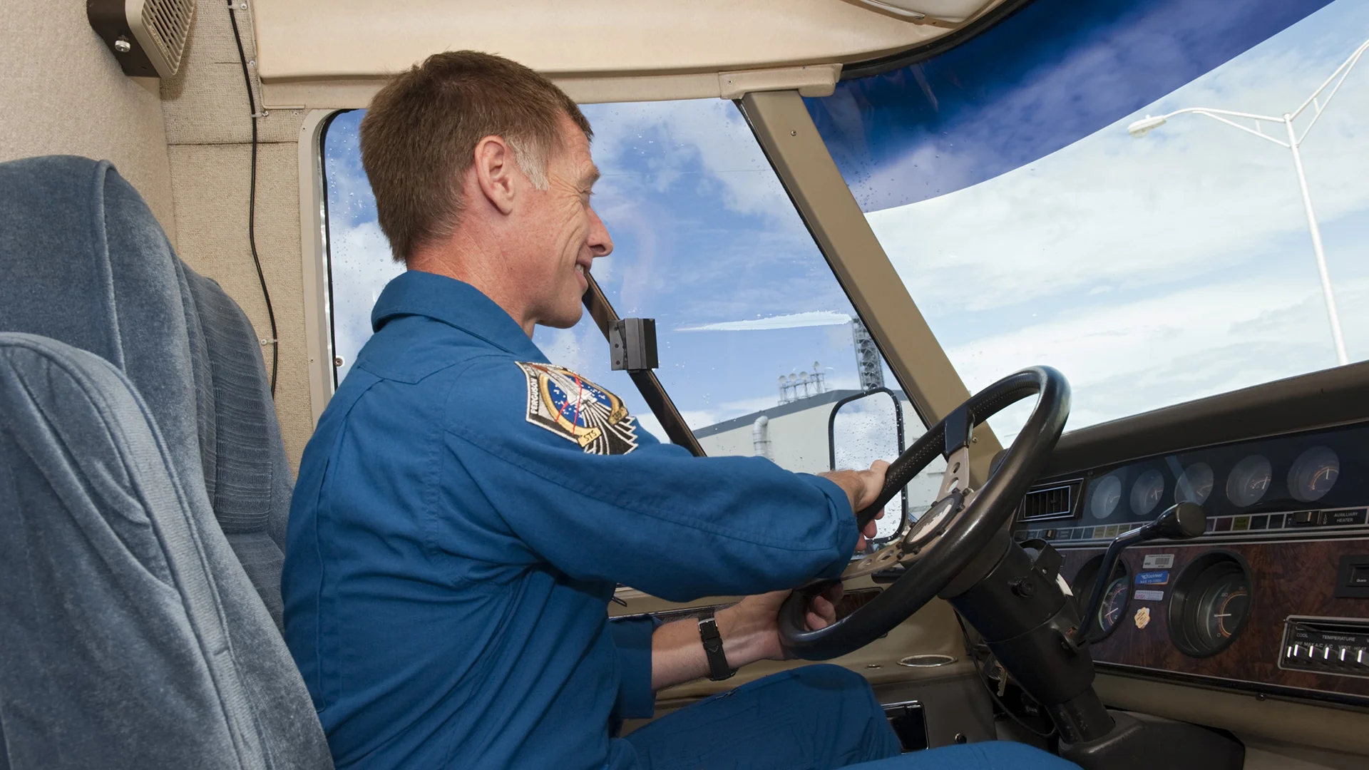Person in a blue jumpsuit driving a vehicle, focused on the road, with a clear blue sky visible through the window.