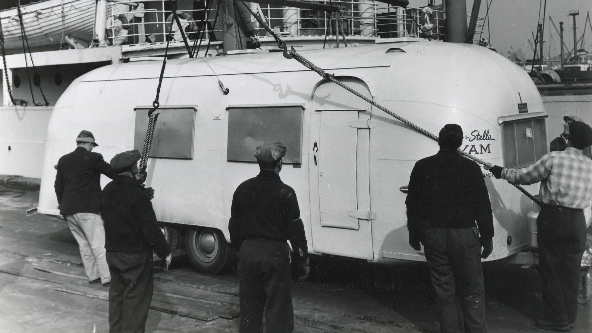 Workers hoist a vintage trailer onto a ship using ropes and cranes.