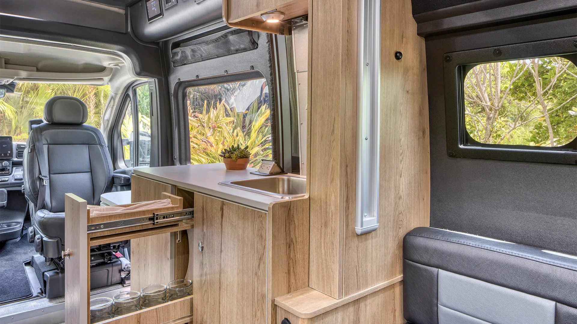 Compact camper van interior with a driver's seat, wooden cabinetry, a pull-out shelf, a small sink, and a potted plant near a window.
