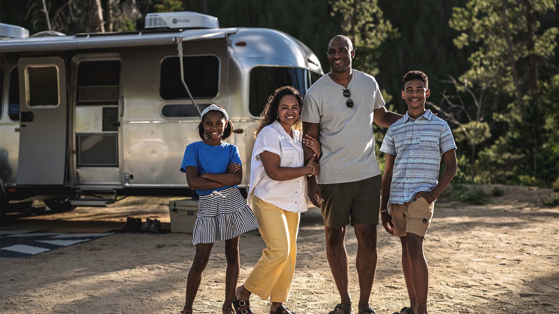 A family of four smiling and posing outdoors in front of a silver camper with trees in the background.