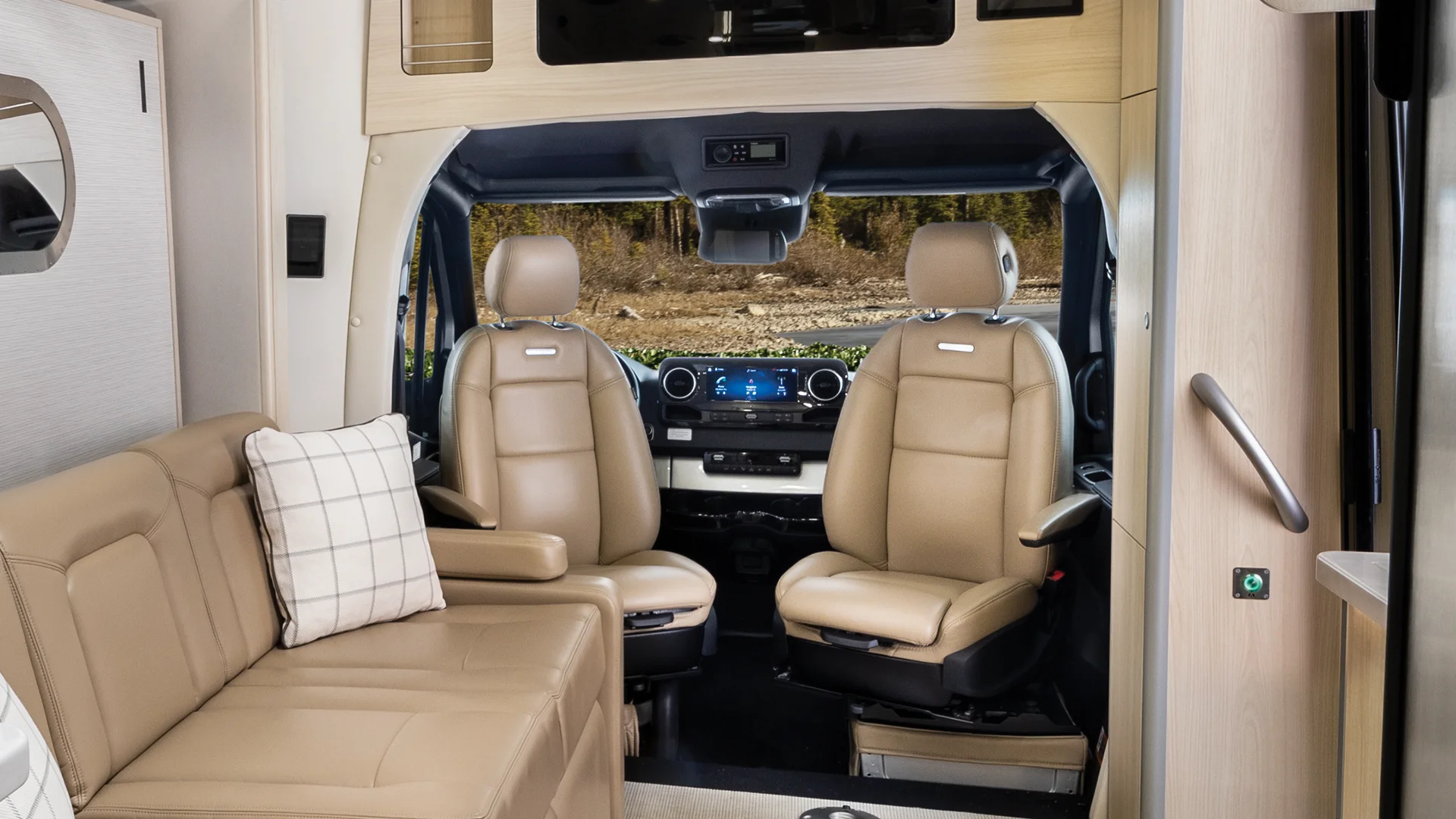 Luxurious RV interior with two leather captain chairs, a plaid cushion on the sofa, and a dashboard facing a scenic forest view outside.