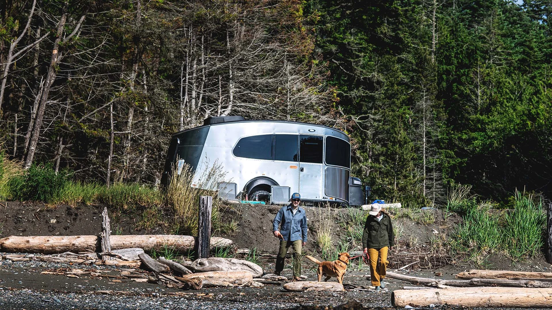 Two people walk a dog on a rocky beach with driftwood, near a shiny silver trailer, surrounded by dense, tall trees.