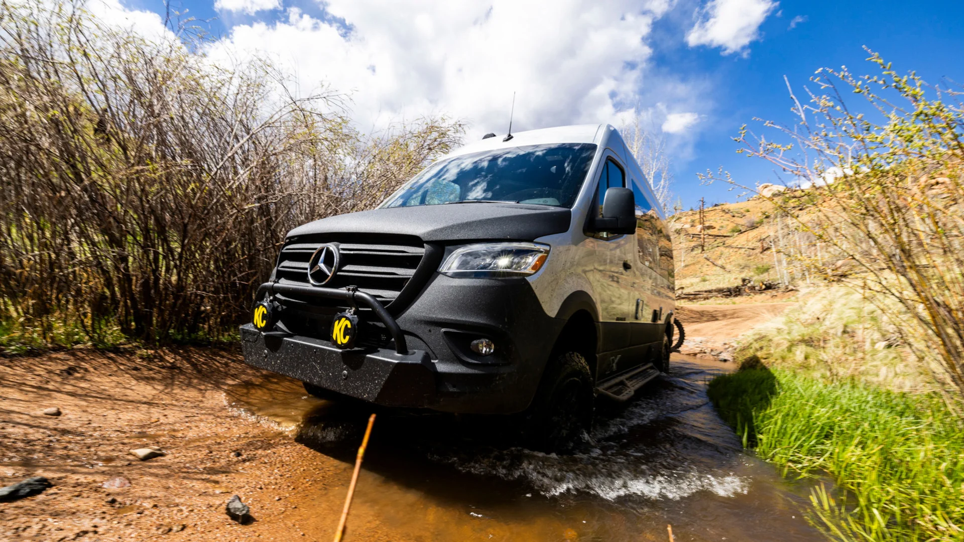 A white van with off-road features crosses a shallow stream on a rugged dirt path, surrounded by tall grass and hills under a partly cloudy sky.