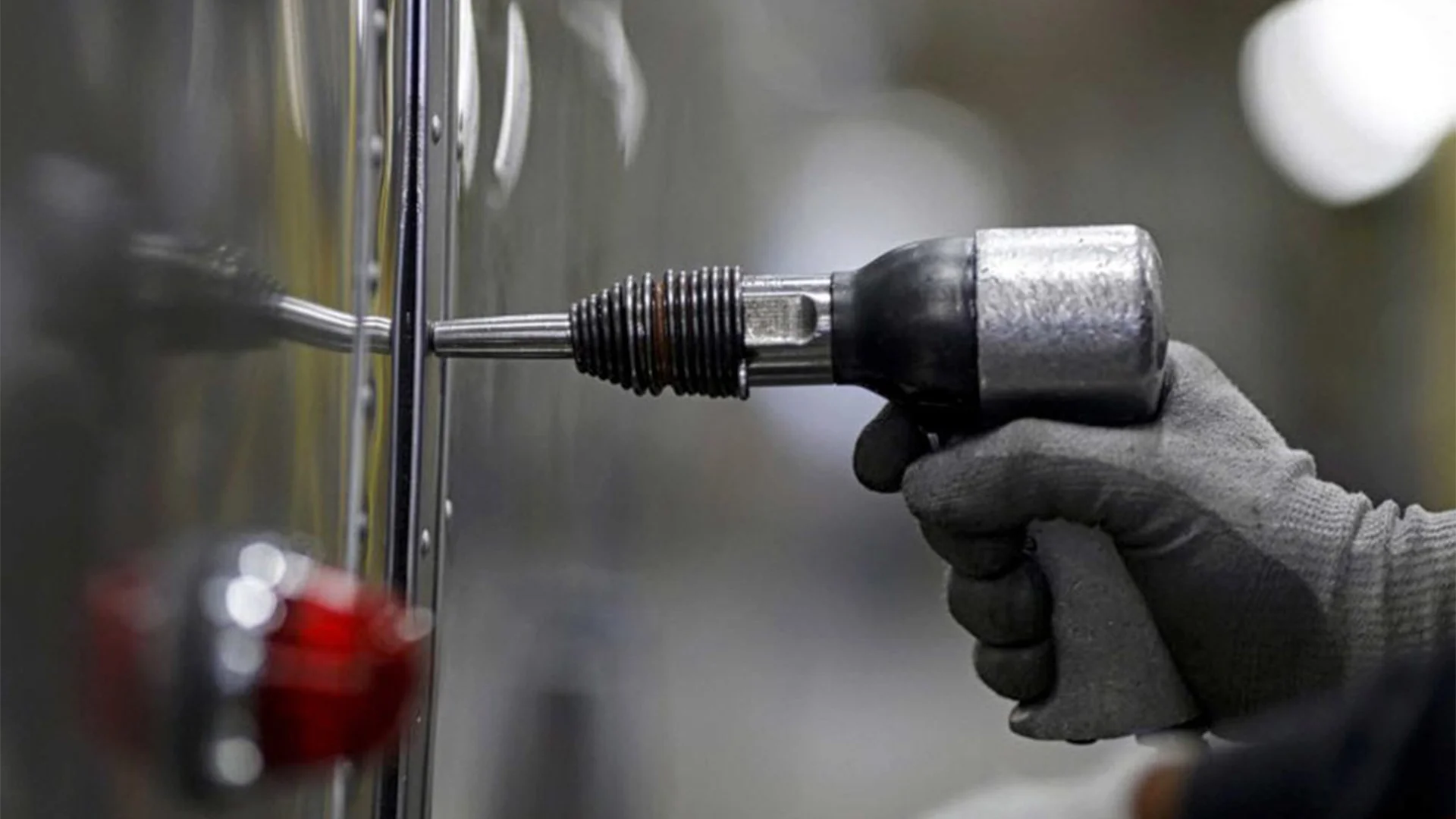 Person wearing gloves uses a pneumatic tool on a metallic surface, with a red knob visible in the foreground.