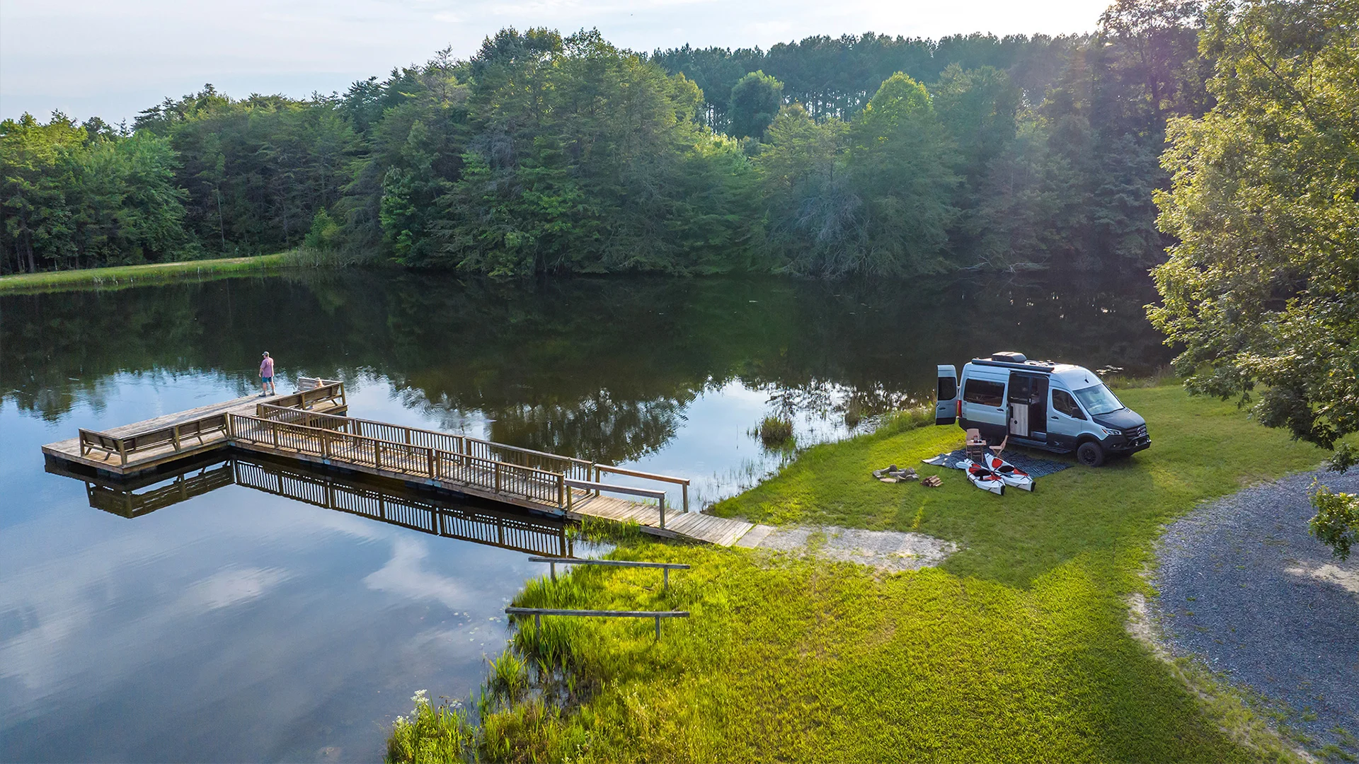 A person stands on a lakeside dock, with a parked camper van, chairs, and equipment nearby on grassy terrain surrounded by trees.