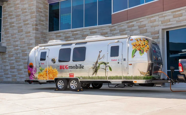 A silver trailer labeled "BUGmobile" with colorful insect and flower illustrations, parked outside a modern brick building attached to a truck.