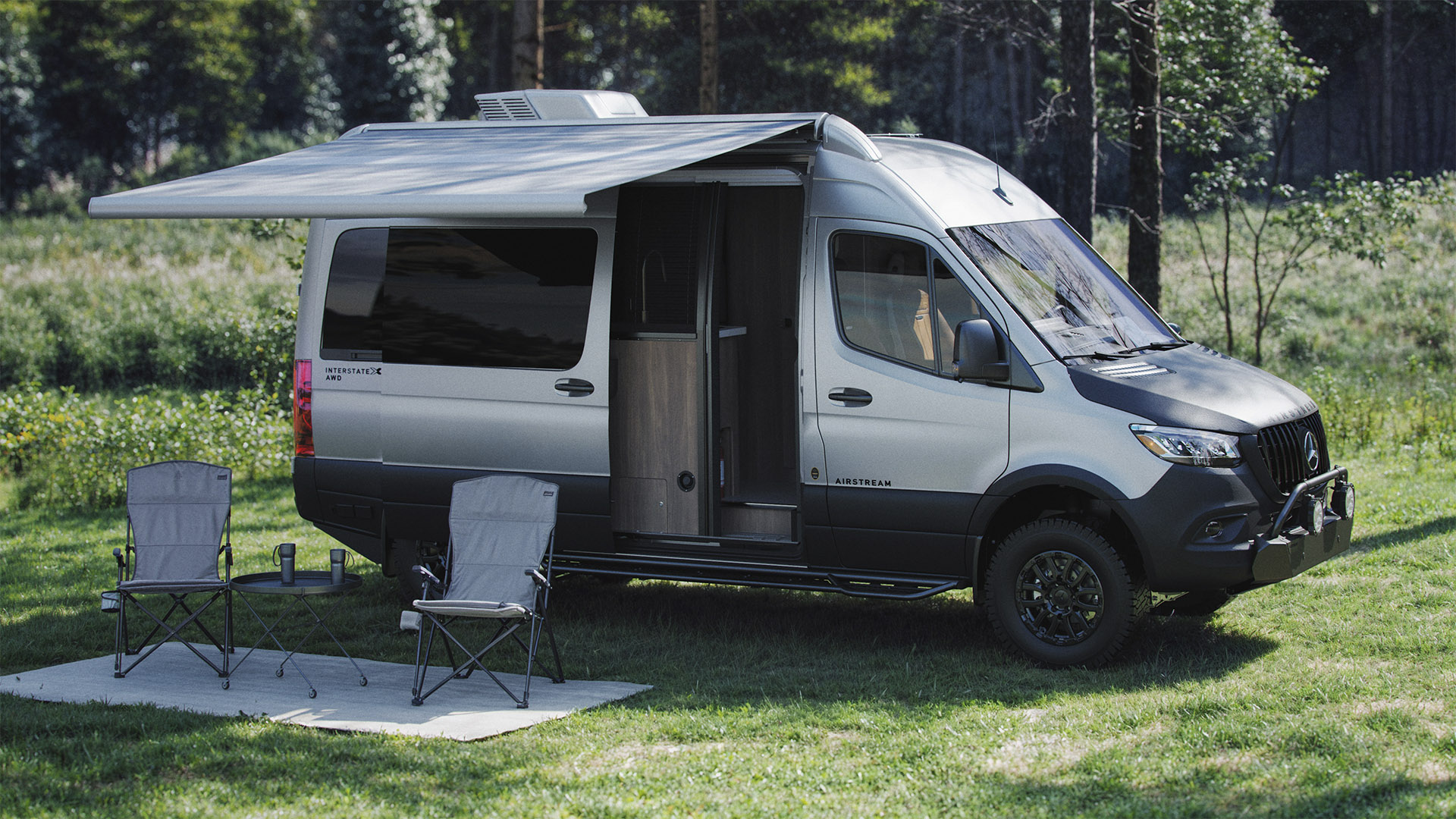 Camper van with extended awning parked on grass, two chairs and a small table nearby, surrounded by trees in a serene setting.