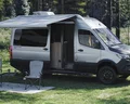 A gray camper van with an extended awning is parked on grass in a scenic outdoor setting. Two folding chairs and a small table are set up on a mat beside the van, surrounded by trees and greenery.