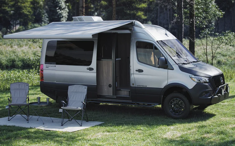 A gray camper van with an extended awning is parked on grass in a scenic outdoor setting. Two folding chairs and a small table are set up on a mat beside the van, surrounded by trees and greenery.