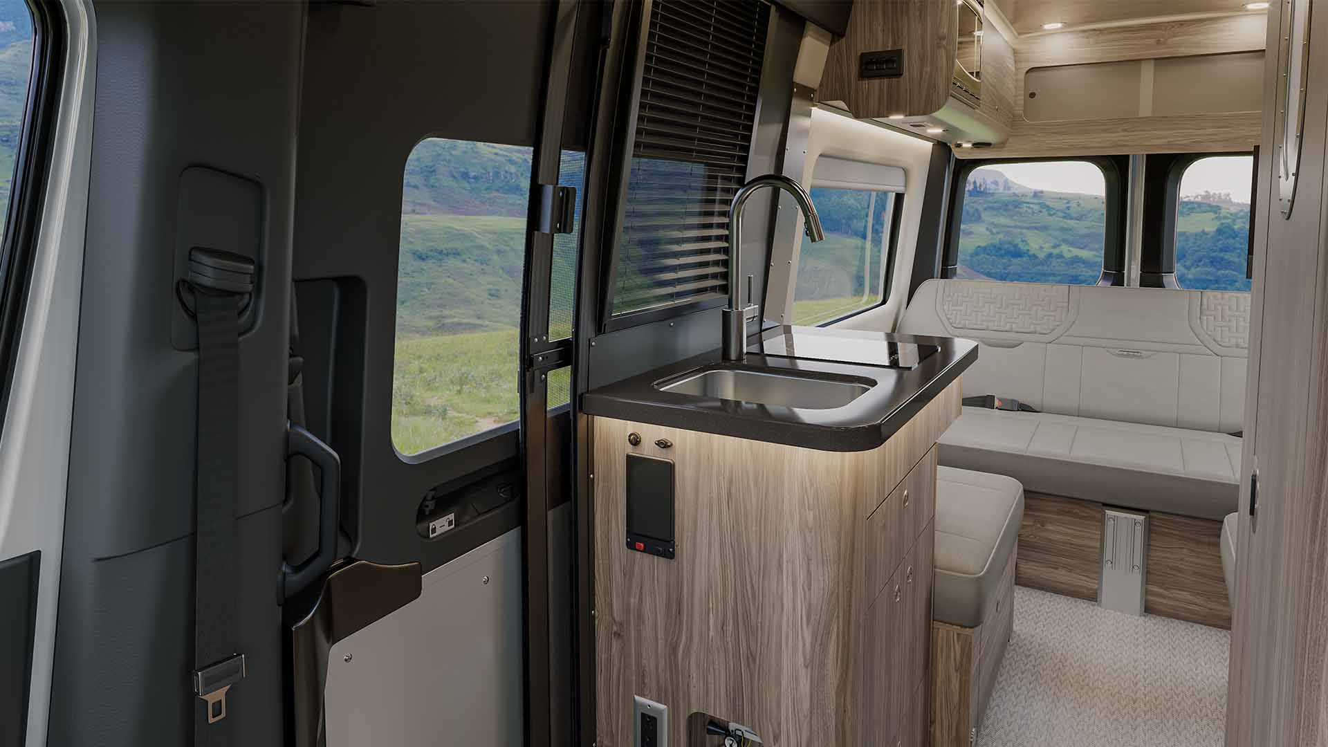 Interior of a modern camper van with wooden cabinets, a sink, and bench seating, set against a scenic landscape view through large windows.