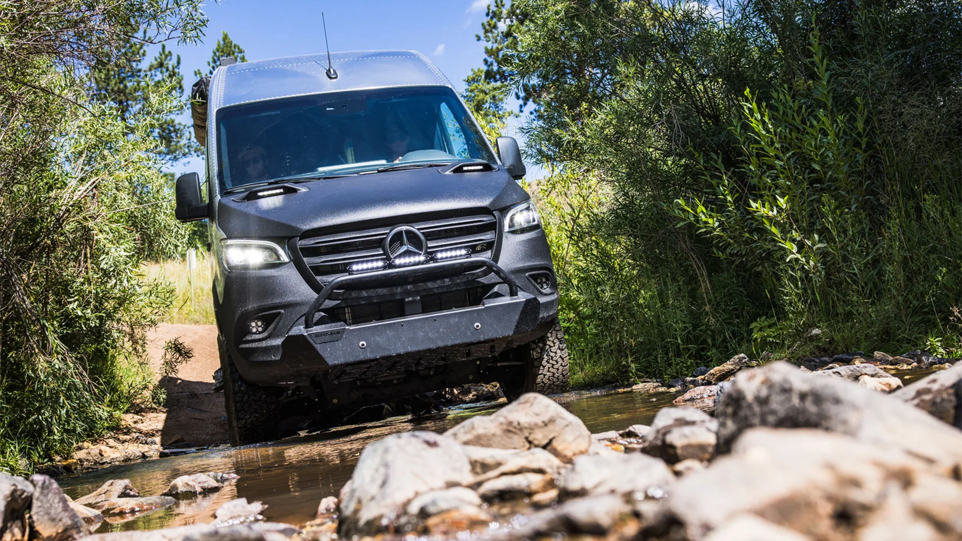 A black Mercedes-Benz van navigates a rocky stream surrounded by lush greenery and trees under a clear blue sky.