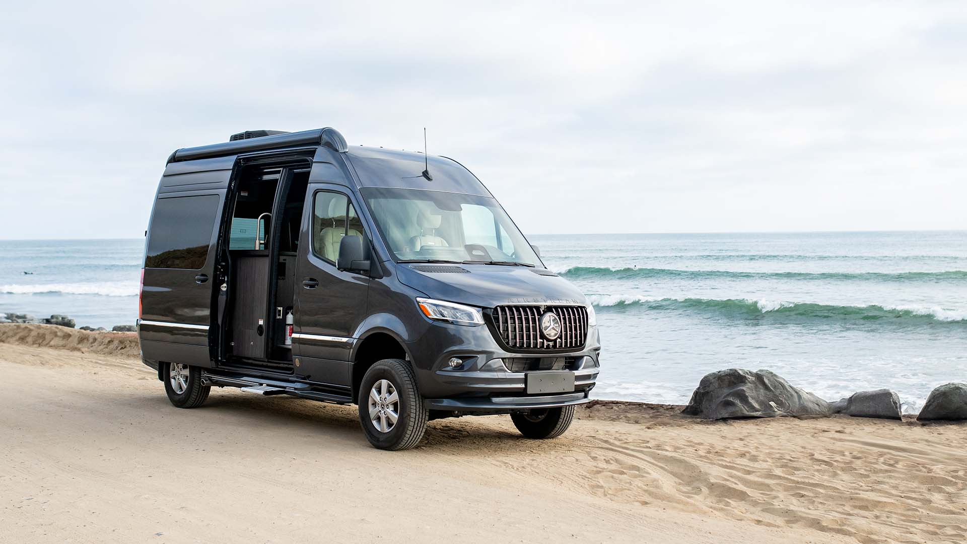 A dark-colored camper van parked on a sandy beach with waves and a cloudy sky in the background.