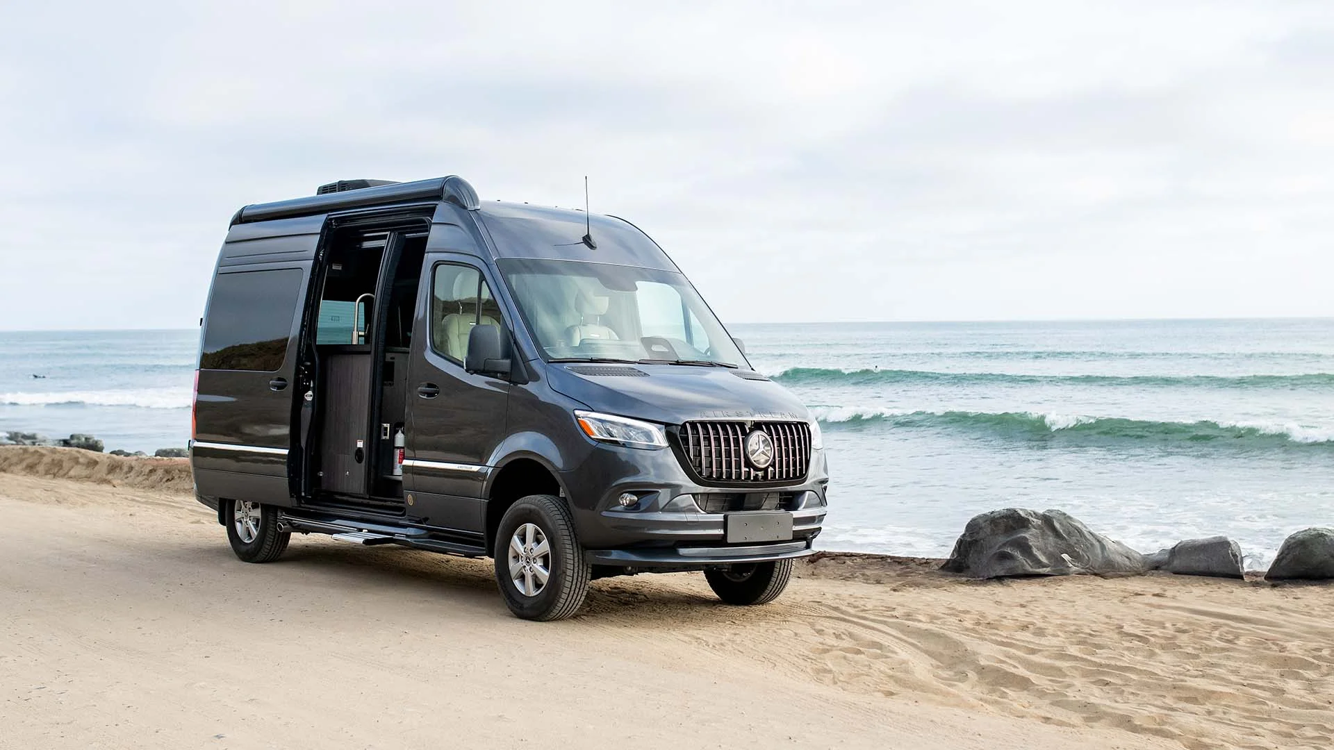 A black camper van parked on a sandy beach with the ocean and cloudy sky in the background.
