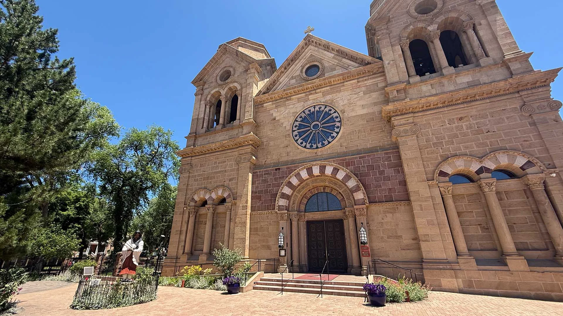 A large stone cathedral exterior with twin bell towers, arched entrance, and a rose window, surrounded by trees and flowers under a clear blue sky.