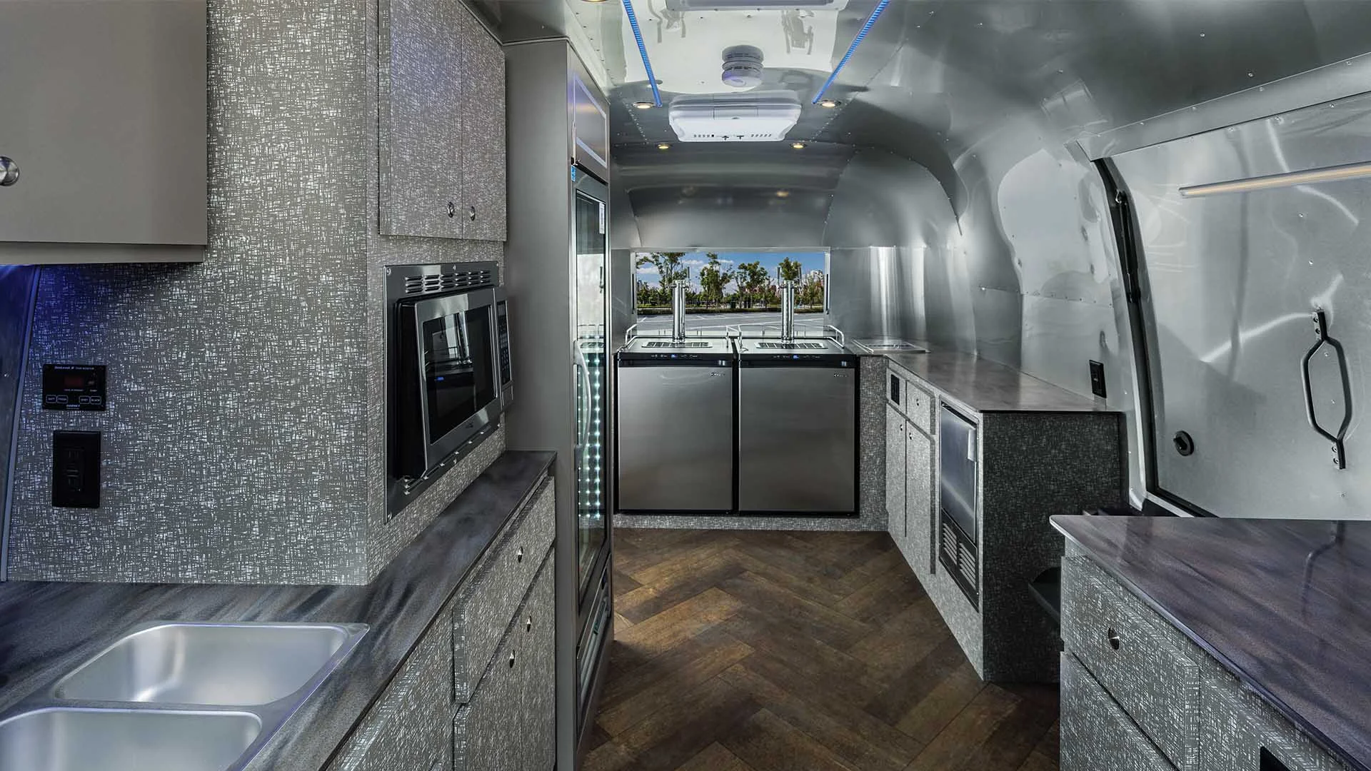 The interior of an Airstream Commercial custom bar and beverage trailer with sleek countertops, built-in microwave, stocked fridge, dual sinks, and overhead storage, featuring ambient ceiling lighting.