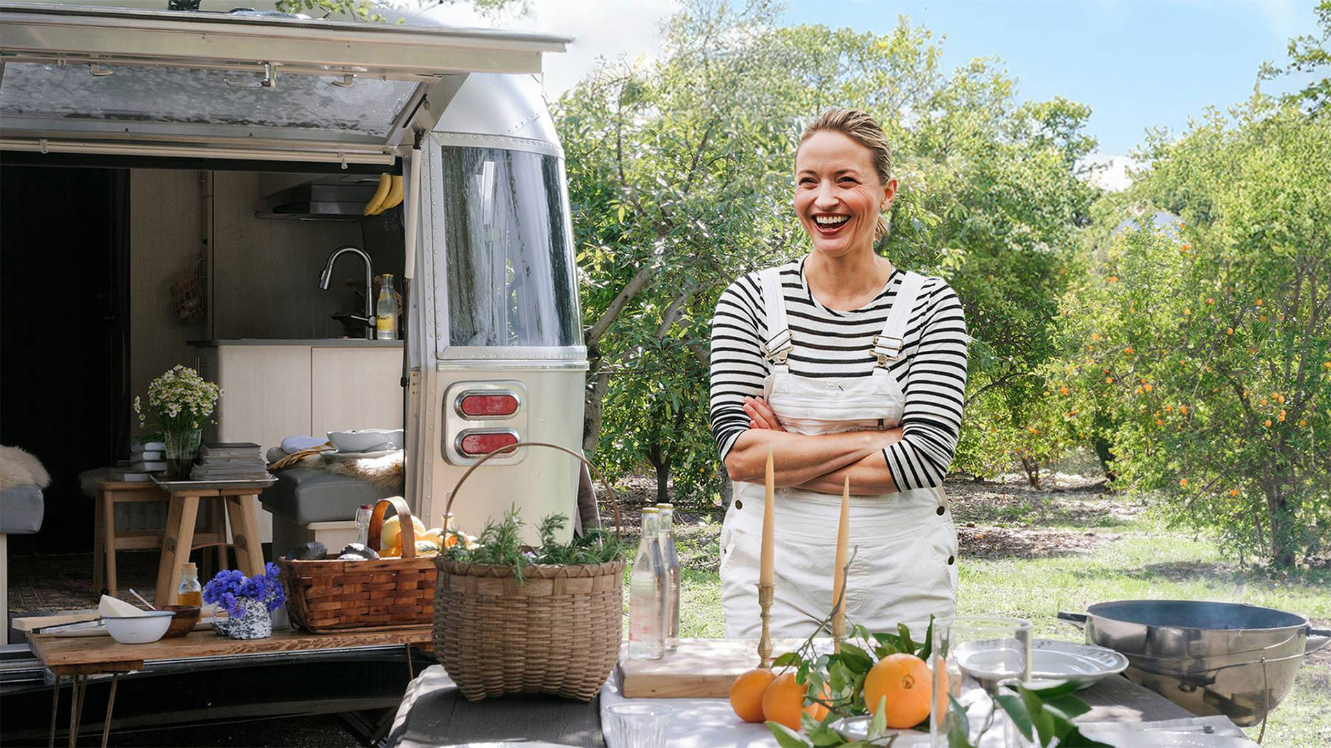 Woman smiling in front of an outdoor kitchen setup, with a vintage trailer in the background and a basket of oranges and flowers on the table.
