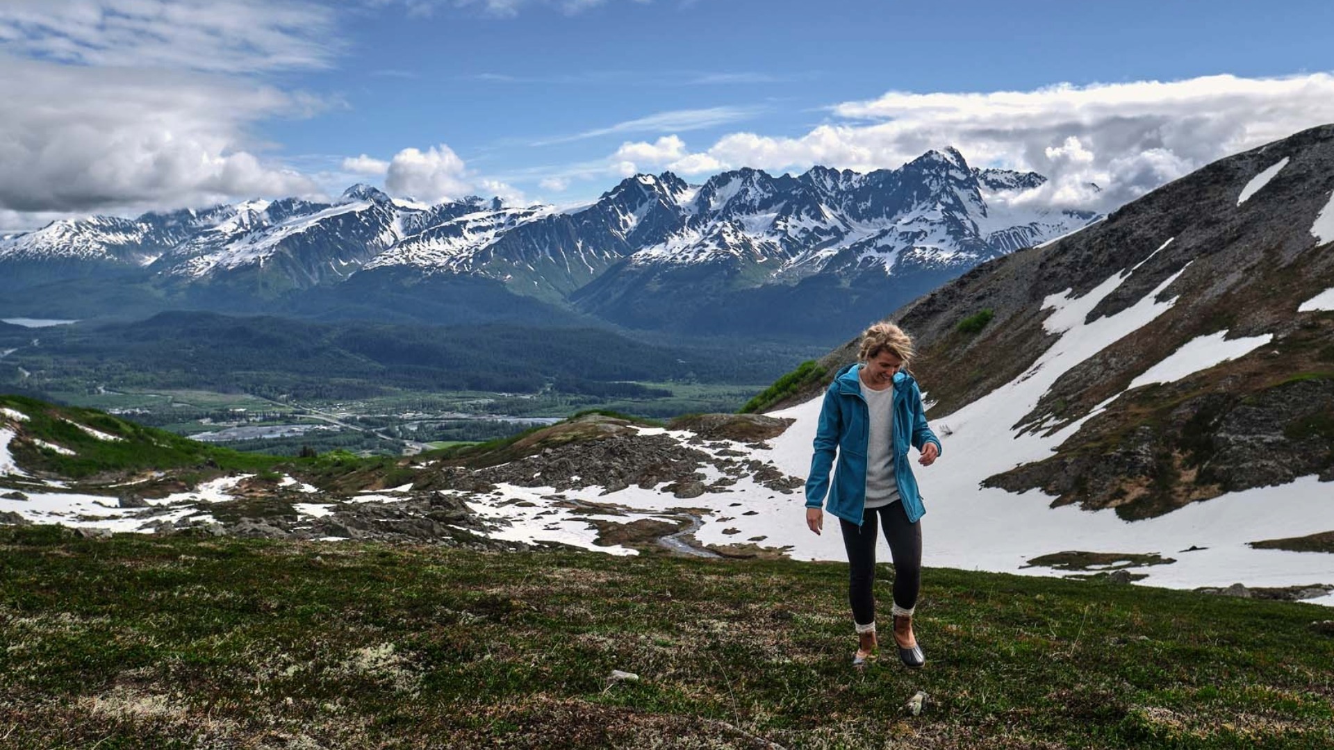 A person in a blue jacket hikes on a grassy mountain with snow patches, with a scenic view of distant snow-capped peaks under a cloudy sky.