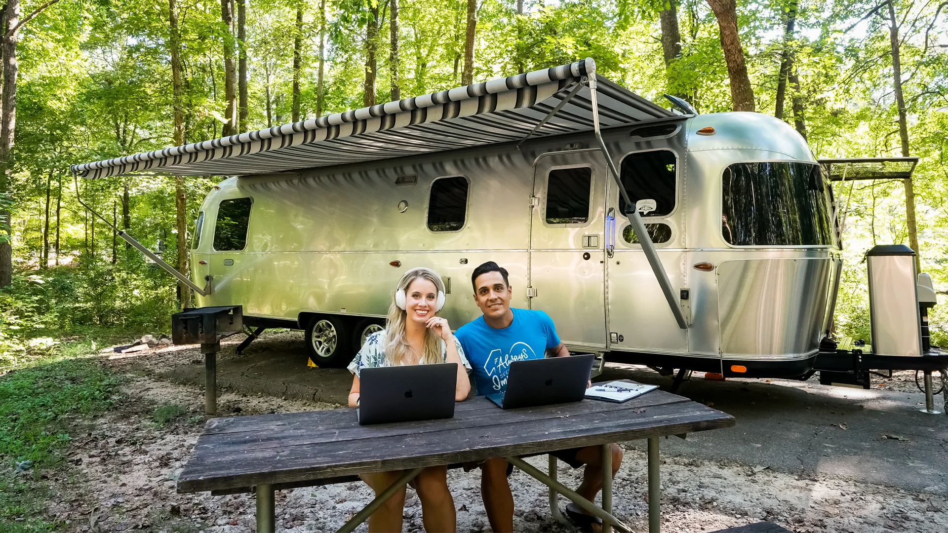 A smiling woman and man sit at a wooden picnic table with laptops, outside a modern silver Airstream trailer parked in a wooded campsite with trees and green foliage all around.