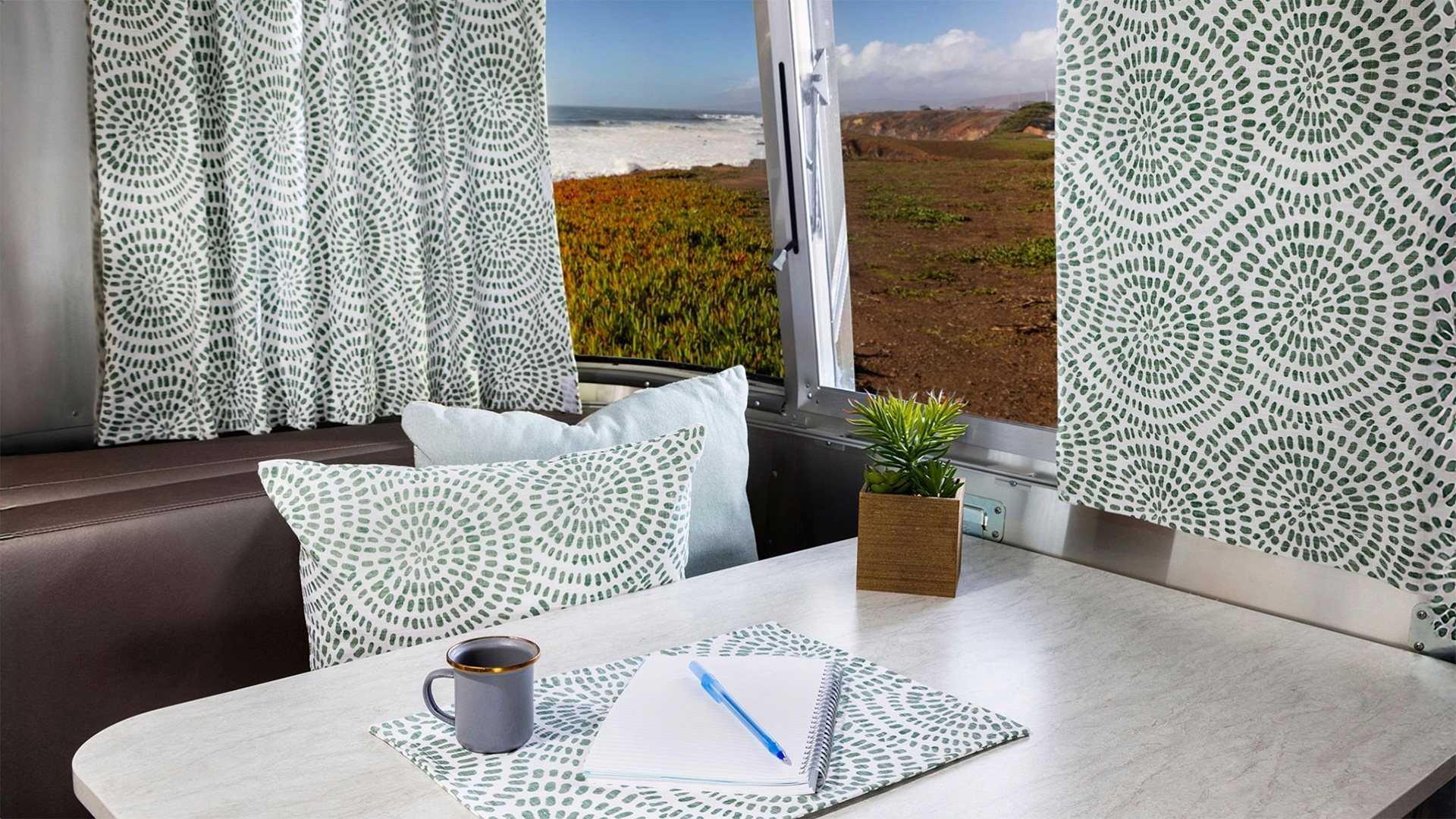 Interior of a camper with green-patterned curtains and cushions, a table set with a notebook, pen, coffee cup, and a small plant, overlooking a coastal view.