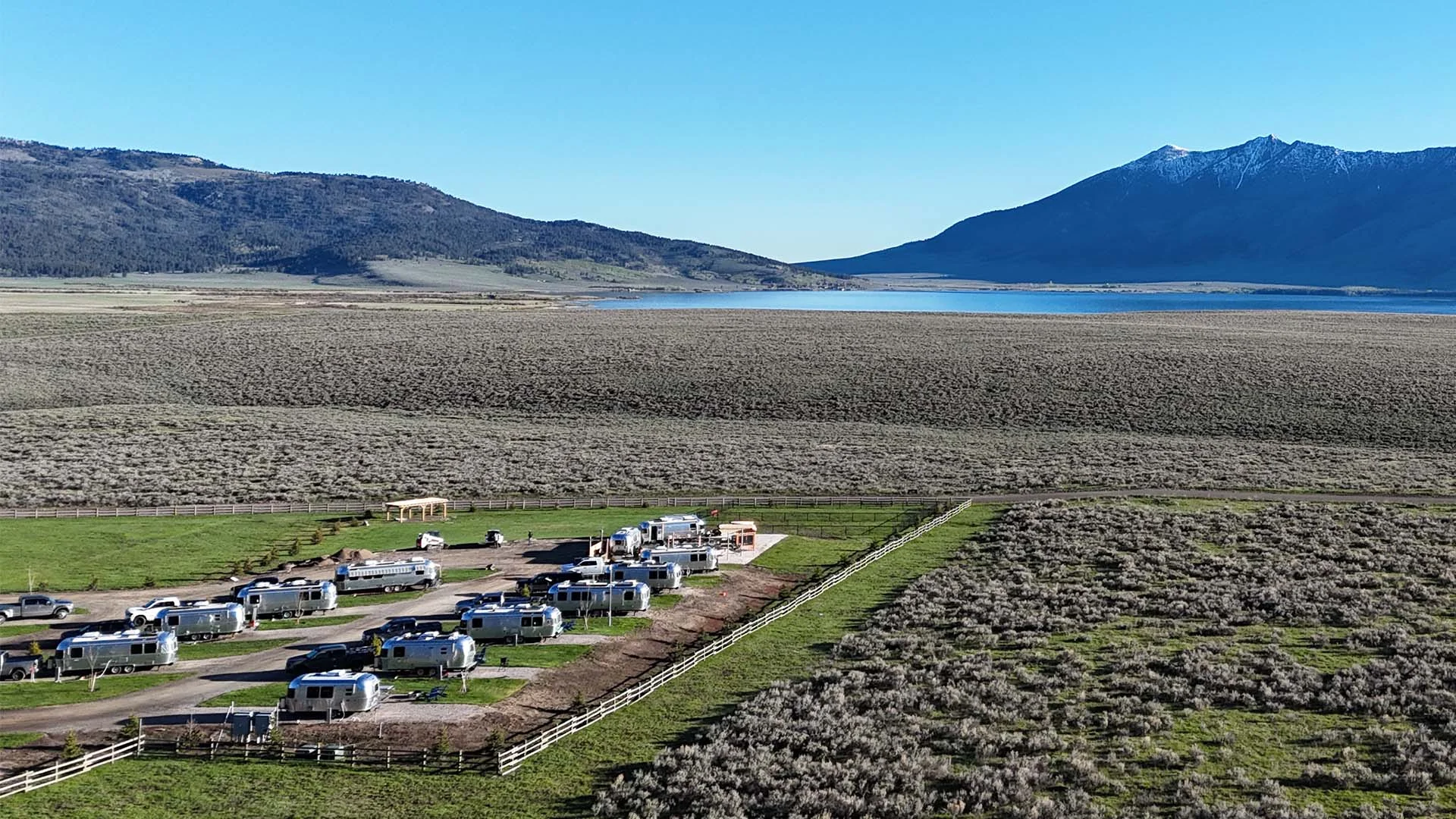 Aerial view of a trailer park with RVs on grassy plots, surrounded by vast fields, mountains, and a distant lake under a clear blue sky.