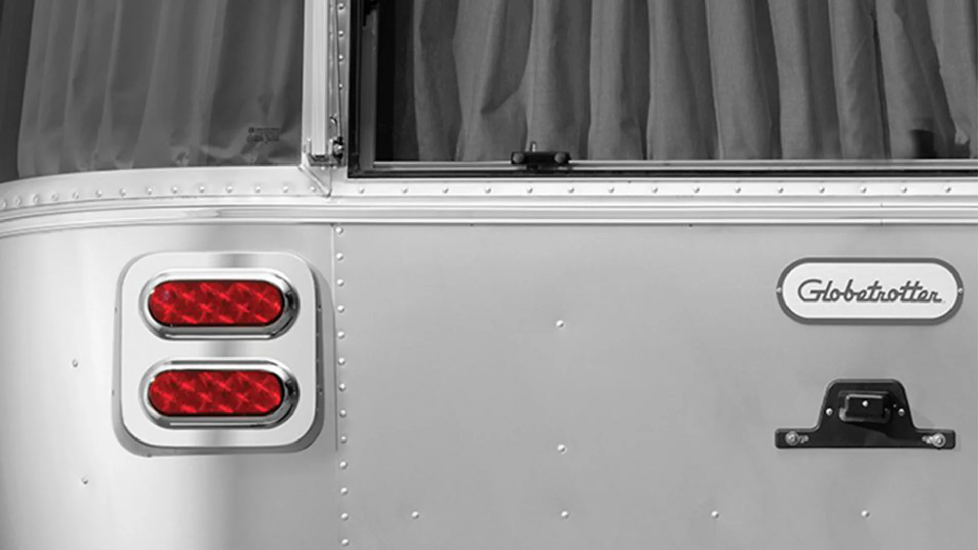 Close-up of a silver trailer with red tail lights, window, and "Globetrotter" label, featuring riveted metal detail and curtain inside.
