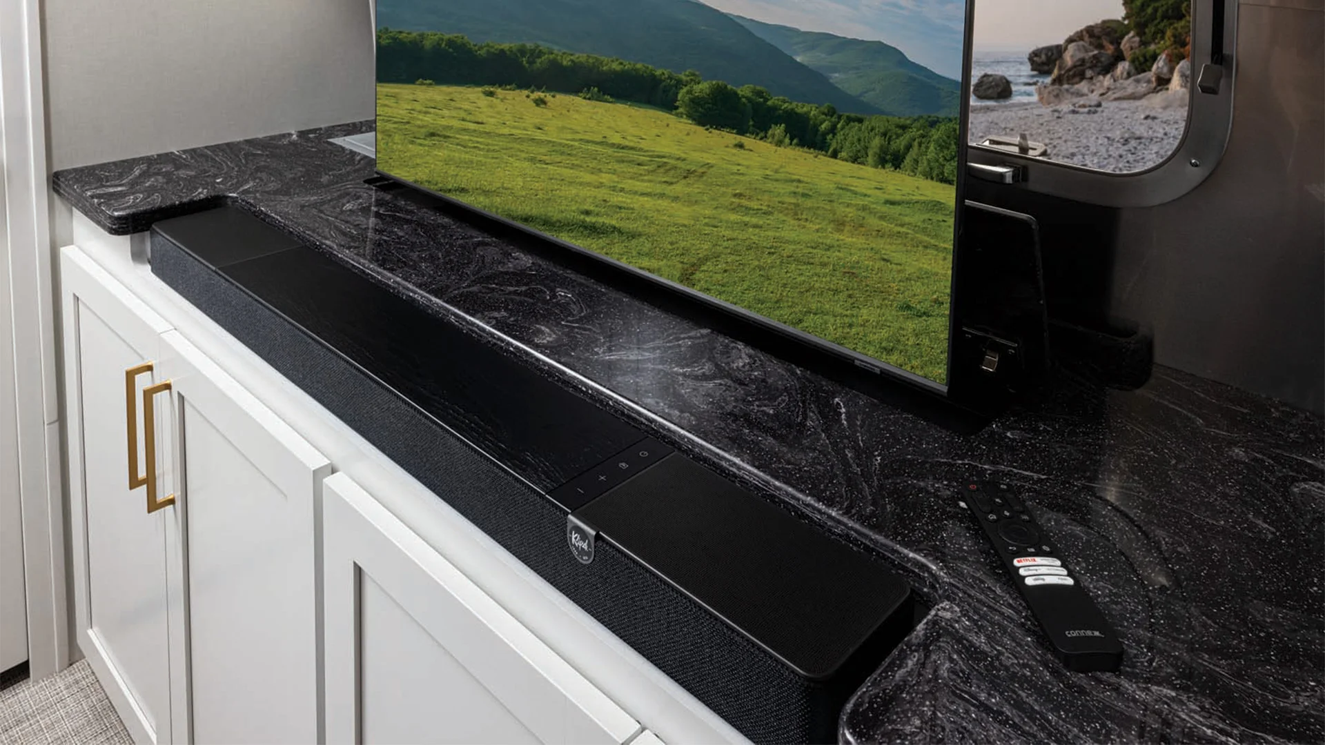 A sleek soundbar on a black marble countertop, in front of a TV displaying a scenic landscape. A remote control is nearby on the counter.