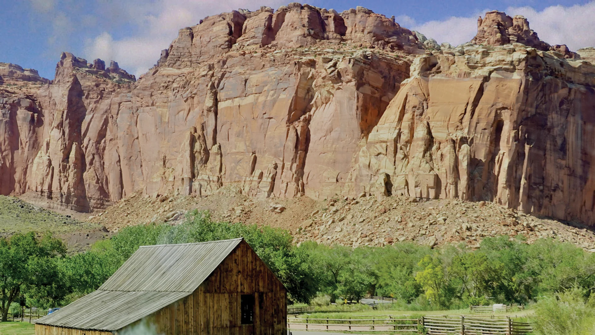A rustic wooden barn sits in a grassy field with trees, backed by towering red rock cliffs under a partly cloudy sky.
